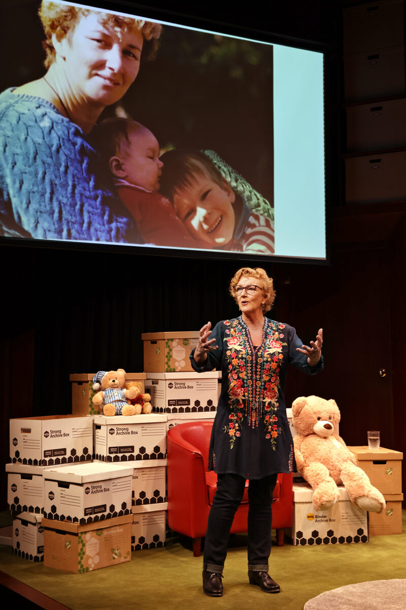 A woman speaks on stage in front of boxes and soft toys, with a projected photo behind her showing a smiling woman holding two small children.