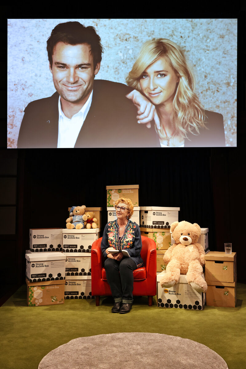 A woman sits on a red chair surrounded by boxes and teddy bears, with a large photo of a smiling man and woman displayed on the screen behind her.