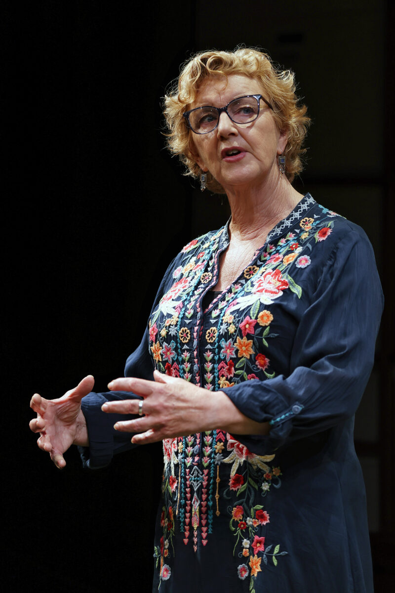 An older woman with short curly hair and glasses speaks passionately, wearing a dark dress with colourful floral embroidery, gesturing with her hands against a dark background.