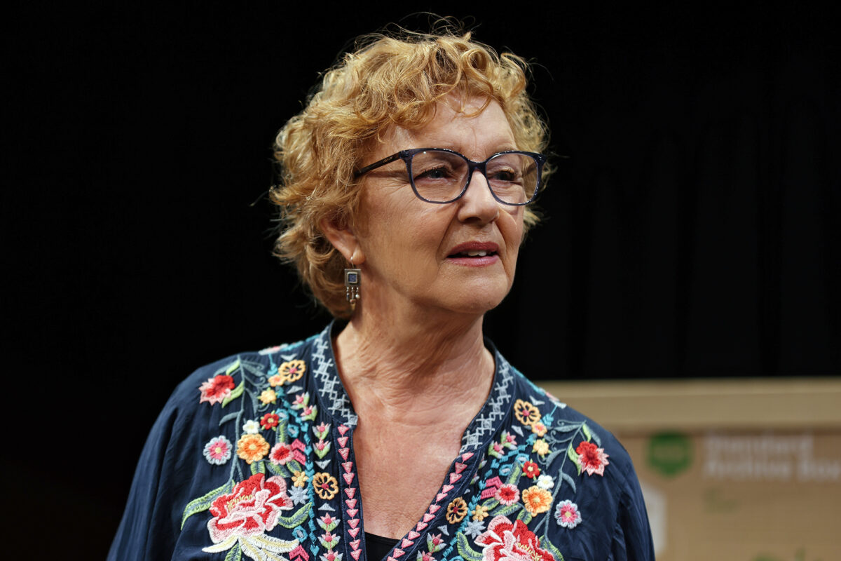 An older woman with curly blonde hair and glasses wears a dark blue top with colourful floral embroidery, standing against a dark background. She has a thoughtful expression and is looking slightly to the side.