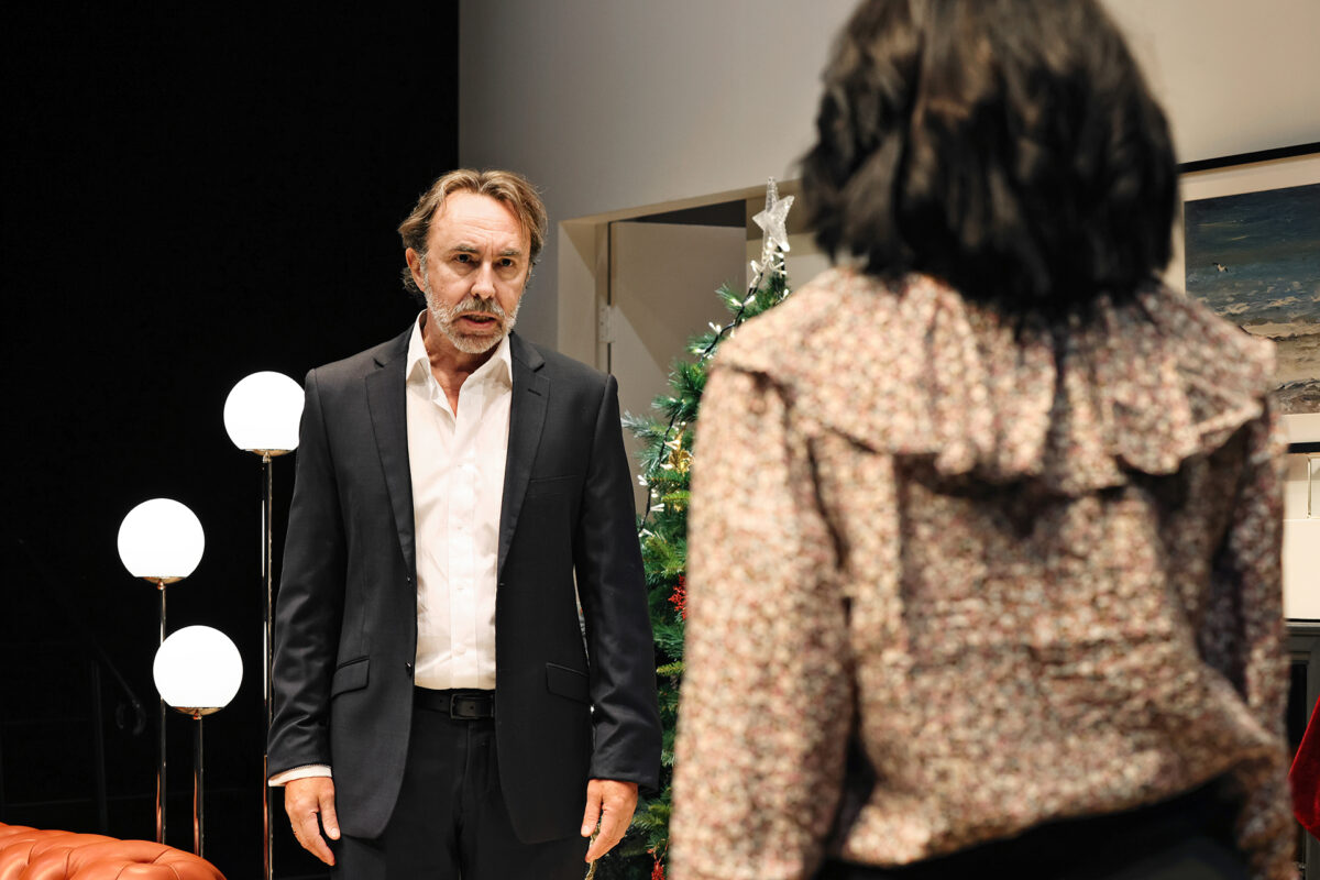 A man in a suit stands with a serious expression facing a woman with shoulder-length dark hair. They are indoors, near a Christmas tree and modern lamps, suggesting a tense conversation.