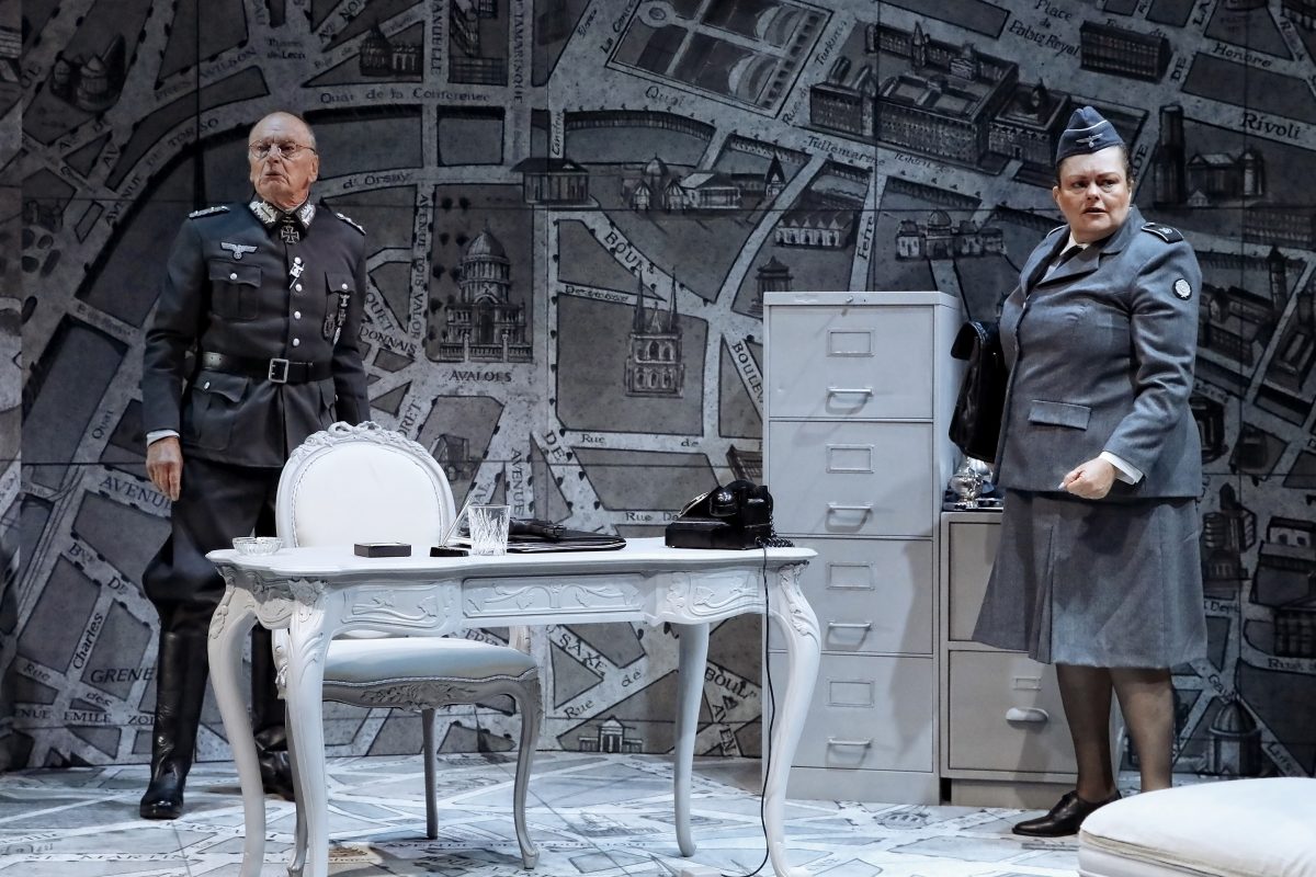 Two people in military uniforms stand in a grey office with a map-covered wall, a white desk with a telephone, and a set of filing cabinets. The setting evokes a Second World War-era command centre.