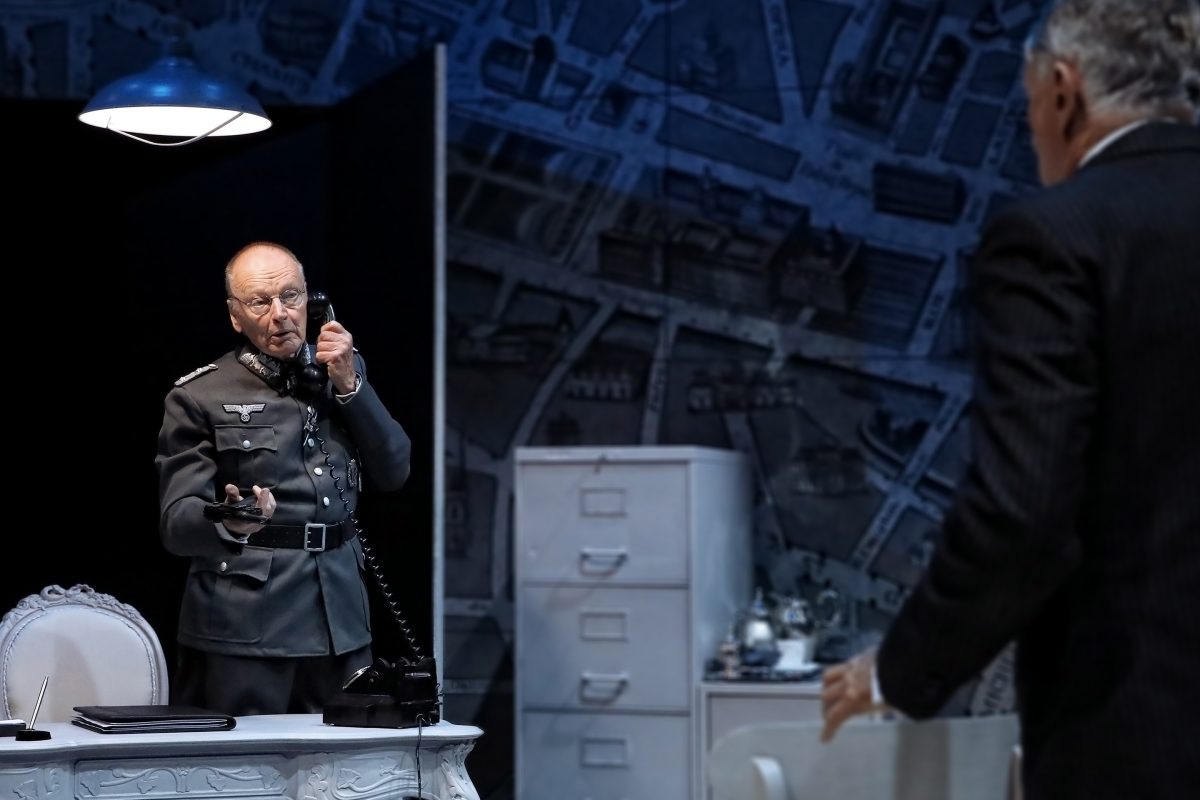 An older man in a WWII-era German military uniform speaks on a vintage telephone at a white desk, whilst another man in a suit stands facing him. The background shows a large map on the wall.