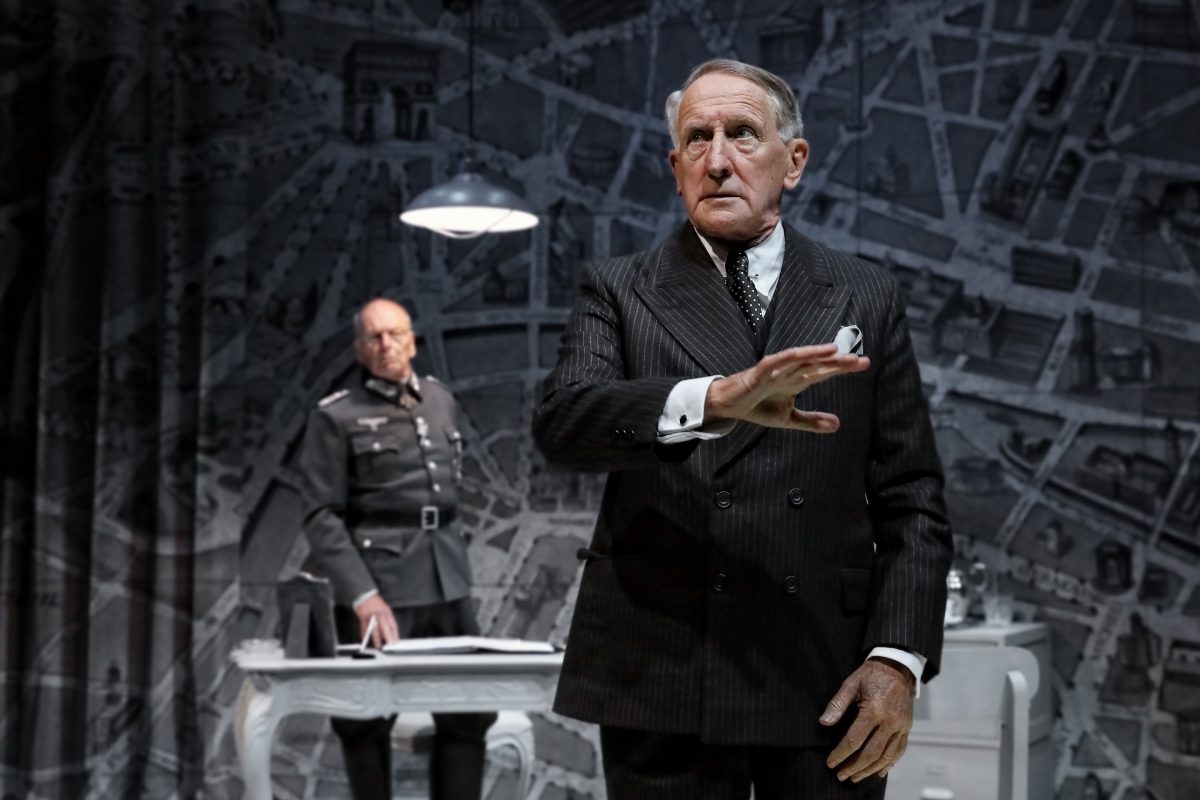 An older man in a dark pinstripe suit gestures whilst speaking, standing in front of a desk. Behind him, a man in military uniform sits at the desk. The background shows a map-like mural.