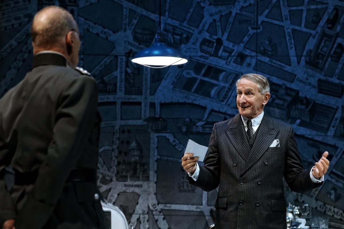 Two men in formal attire converse on stage; one wears a dark suit and holds a paper, gesturing expressively, while the other, seen from behind, wears a uniform. A large map is displayed as the backdrop, with a lamp above them.