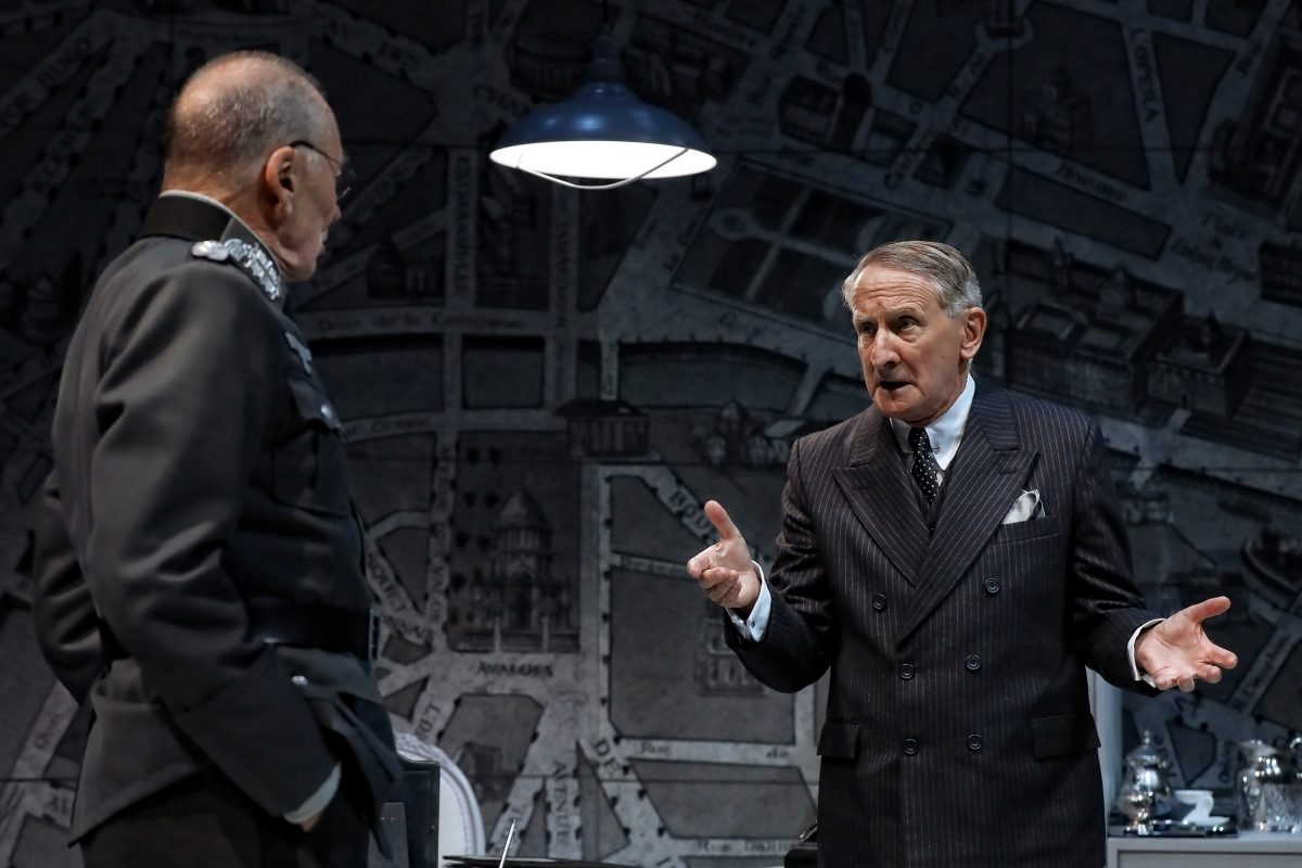 Two men in formal attire, one in a military uniform and the other in a striped suit, are having an intense conversation under a hanging lamp, with a large city map in the background.