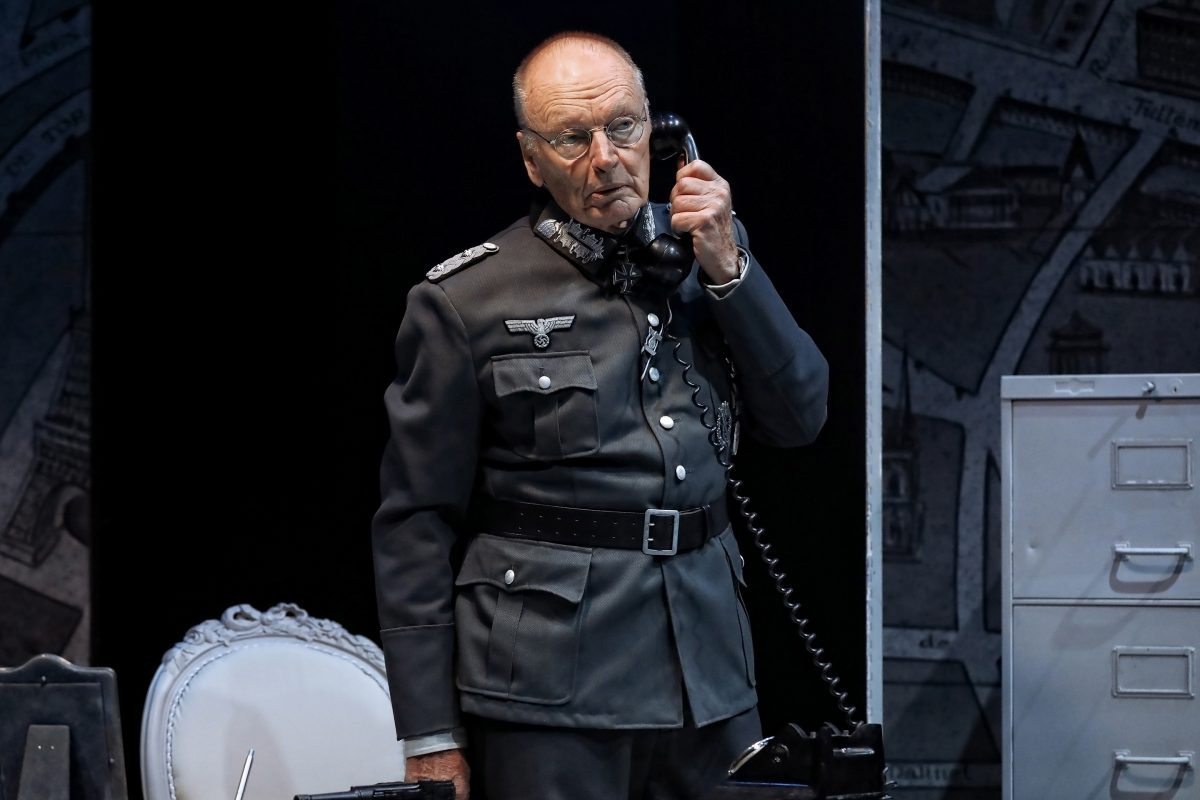 An older man in a grey military uniform with Nazi insignia stands indoors, holding a black telephone receiver to his ear. He appears serious, and office furniture is visible around him.