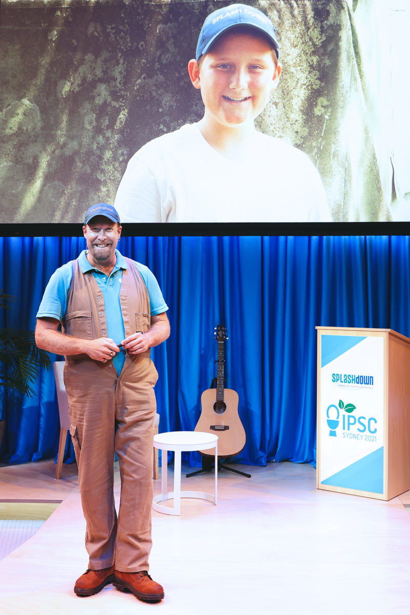 A man in khaki overalls and a blue cap stands on a stage with a guitar, a small table, and a lectern labelled IPSC Sydney 2021. Behind him is a large screen showing a smiling boy in a cap.