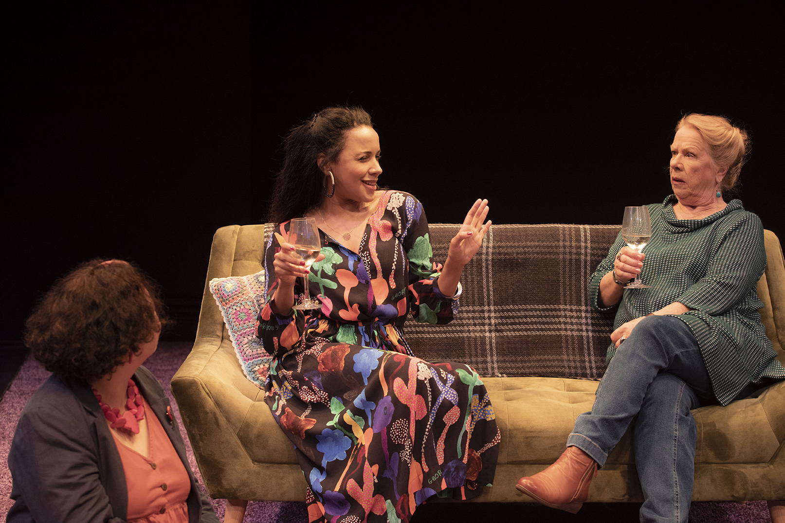 Three women sit on a sofa talking. The woman in the centre gestures with her hand while holding a wine glass; the woman on the right looks surprised and also holds a glass. The woman on the left looks towards them.