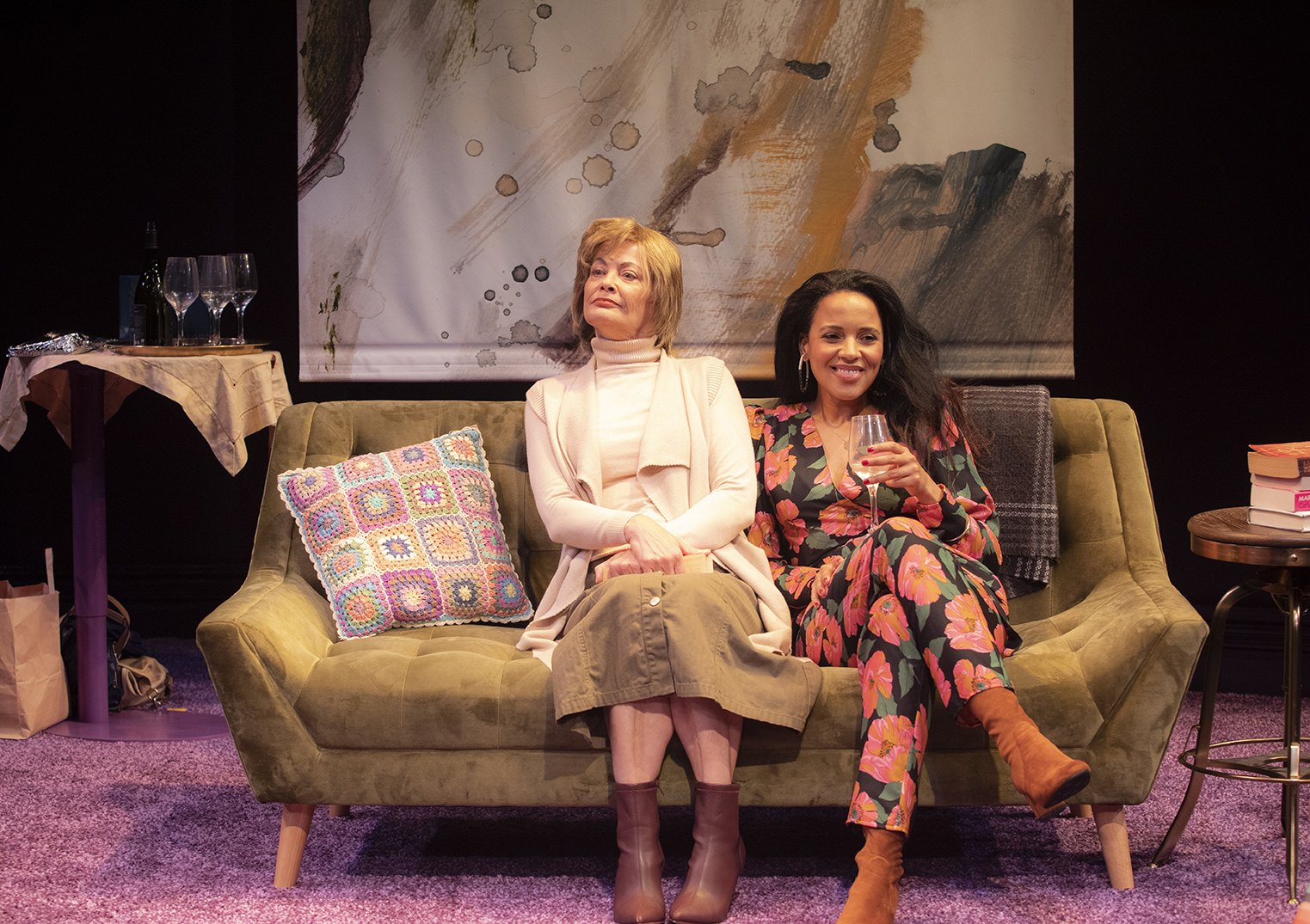 Two women sit on a green sofa in a cosy room. One wears a light polo neck and skirt, looking calm; the other wears a colourful floral outfit and smiles, holding a wine glass. Art and glasses are on a table nearby.