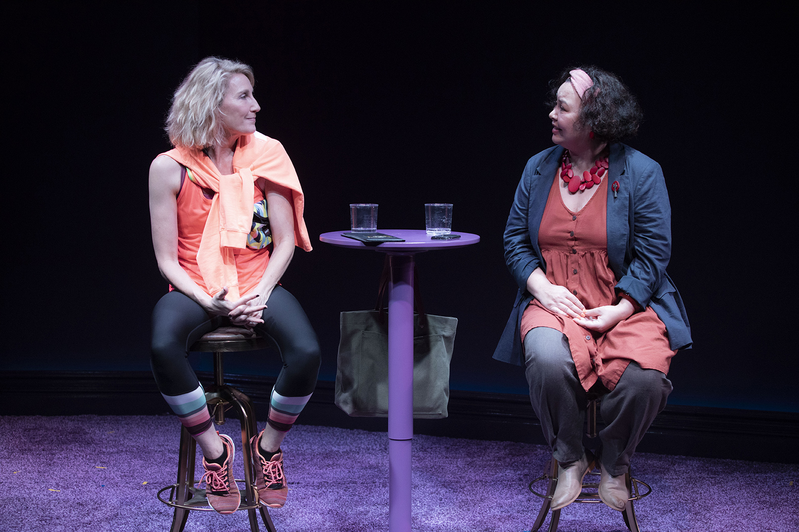 Two women sit on stools at a small round table with two glasses of water. One woman wears sports clothes, the other wears a dress and jacket. They face each other and appear to be having a conversation on stage.
