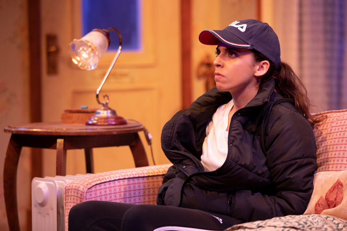 A woman in a dark jacket and baseball cap sits on a patterned sofa, looking thoughtful. A lit table lamp and door are visible in the warmly lit background, creating a cosy indoor setting.