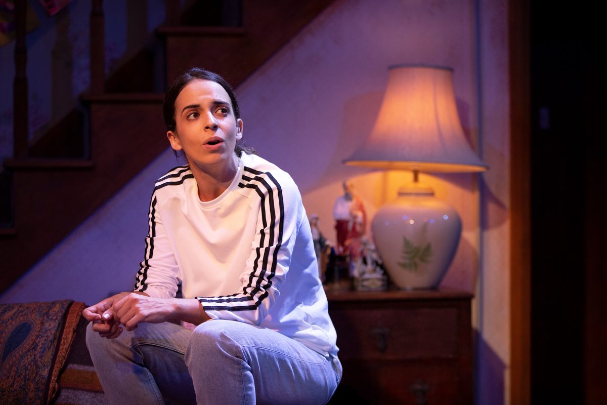 A woman in a white shirt with black stripes sits on a sofa, looking to the side with a thoughtful expression. Behind her, there is a lit lamp, a staircase, and a small table with figurines.