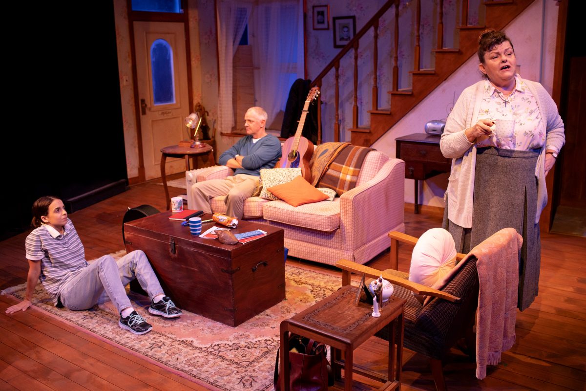 Three people in a cosy living room: an older man sits on a sofa, a younger person sits on the floor by a coffee table, and an older woman stands talking, gesturing with her hand, near a staircase in the background.