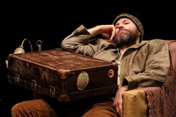 A bearded man wearing a knitted hat and casual clothes lounges on a chair, resting his head on one hand. An old, weathered suitcase sits on his lap, and a textured blanket covers the armrest. The background is dark.
