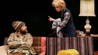 A woman with blonde hair angrily points her finger at a bearded man in a knitted hat sitting with arms crossed on a striped sofa; a lamp and blankets are in the background.