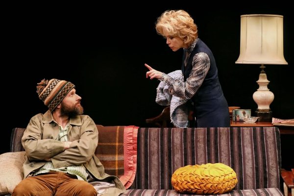 A woman with blonde hair angrily points her finger at a bearded man in a knitted hat sitting with arms crossed on a striped sofa; a lamp and blankets are in the background.