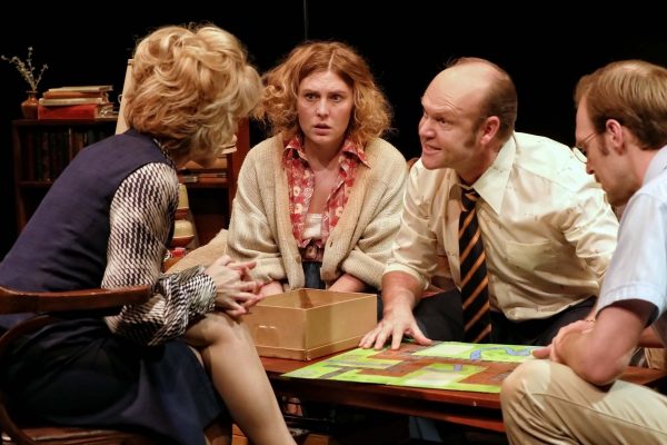 Four adults sit around a table playing a board game. One woman looks concerned, another listens intently, while a man in a striped tie leans forward, speaking passionately. A bookcase is in the background.