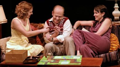 Three adults sit on a sofa, laughing and interacting while playing a board game on a coffee table in front of them. They appear relaxed and joyful, surrounded by cosy lamps and cushions.