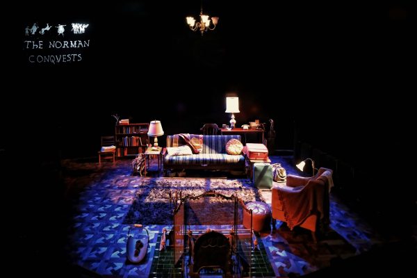 A warmly lit stage set depicts a cosy living room with a sofa, armchairs, bookshelves, lamps, and rugs. The text The Norman Conquests is displayed in the upper left corner against a dark background.