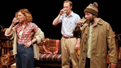 Three people stand in front of a sofa, each drinking from a glass. A woman in a cardigan and patterned shirt is on the left, a man in glasses and a tucked-in shirt in the centre, and another man in a beanie and coat on the right.