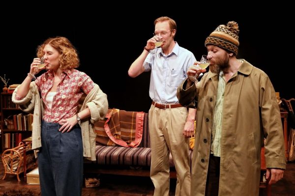 Three people stand in front of a sofa, each drinking from a glass. A woman in a cardigan and patterned shirt is on the left, a man in glasses and a tucked-in shirt in the centre, and another man in a beanie and coat on the right.