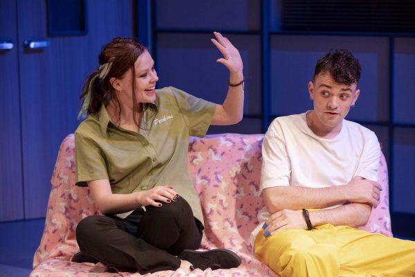 A woman in a green shirt with Phoebe embroidered on it gestures animatedly whilst sitting on a pink patterned sofa next to a man in a white shirt and yellow trousers, who looks uncertain.