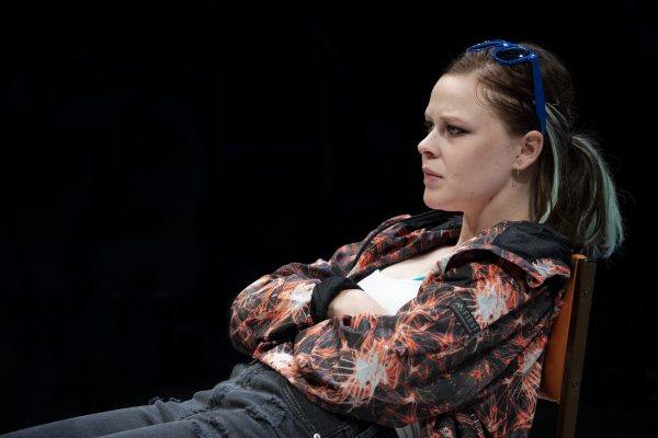 A young woman with fair hair tied back, wearing blue sunglasses on her head, a patterned jacket, and ripped black jeans, sits with her arms folded and a serious expression, looking to the side.