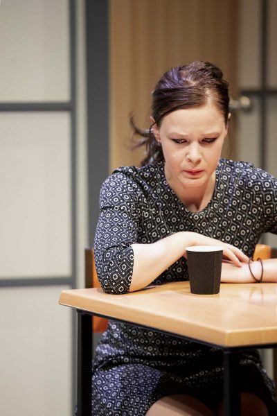 A woman with dark hair, wearing a patterned dress, sits at a table and looks down thoughtfully at a black paper cup in front of her.