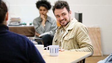 A man in a tan jacket smiles whilst sitting at a table with mugs, as a woman in the background sits with her hand on her chin, watching the conversation.