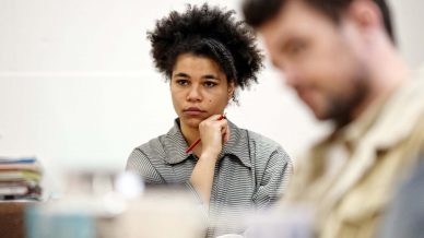 A person with curly hair and a striped shirt sits thoughtfully with a red pen at their chin, whilst another person in the foreground appears out of focus.