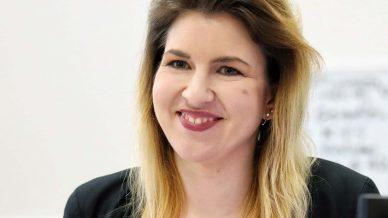 A woman with long, blonde hair and a black blazer smiles while sitting at a desk, with a blurred white background and some text visible behind her.