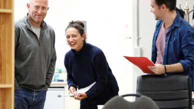 Three people are standing and smiling in an office setting. A bald man in a grey jumper stands to the left, a woman in a dark jumper is laughing in the middle, and a man in a blue shirt holds a red folder on the right.