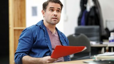 A man with short dark hair, wearing a blue shirt over a mauve T-shirt, sits at a table holding a red folder. He looks slightly upwards with a focused expression. Office furniture and shelves are visible in the background.