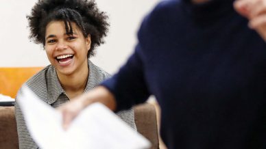 A person sits on a sofa, smiling and laughing, while another person in the foreground gestures energetically, holding papers. The scene appears lively and engaging.