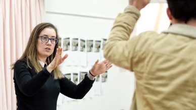 A woman with glasses gestures with her hands whilst speaking to a person in a tan jacket, who raises one hand. They are indoors with a pink curtain and photos on the wall in the background.