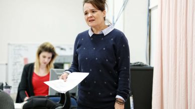 A woman holding papers stands and speaks with a concerned expression in an office. Another person sits and works at a desk in the background, partially out of focus.
