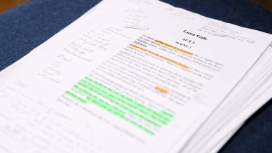 A stack of printed script pages with handwritten notes in the margins. Some lines of the text are highlighted in green and orange. The pages are resting on a dark blue fabric surface.