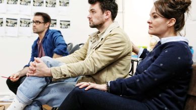 Three people sit closely together, facing forward and listening attentively. Papers and notes are pinned to the wall behind them, suggesting a meeting or workshop setting.