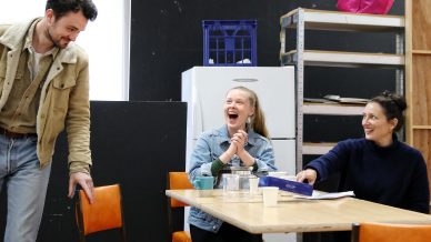 Three people are gathered around a table, smiling and laughing. One man stands by a chair, while two women sit at the table, which has cups and a teapot. The background includes shelves and a fridge.
