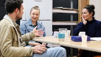 Three people sit around a table in conversation. Two women and one man are engaged, with coffee cups, a teapot, and a box of tissues on the table. The setting appears casual, possibly a break room or informal meeting space.