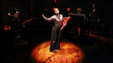 A woman stands in a spotlight, holding an umbrella and surrounded by falling rose petals. Musicians play a cello, piano, and sing in the dimly lit background. The stage floor is covered with rose petals.