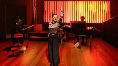 A woman strikes a dramatic pose on a stage with warm lighting. Behind her, a pianist plays a grand piano and a cellist plays a cello. The background features a modern, glowing, wavy panel.