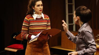 Two women are talking intensely indoors. The woman on the left, holding a folder, looks concerned and wears a colourful striped jumper and brown skirt. The woman on the right gestures with her hand while speaking.