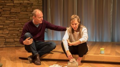 A man and woman sit on a wooden stage. The man gently rests his hand on the woman’s shoulder as she looks down, sorting through letters and papers spread in front of her. A stone wall and soft curtains are in the background.