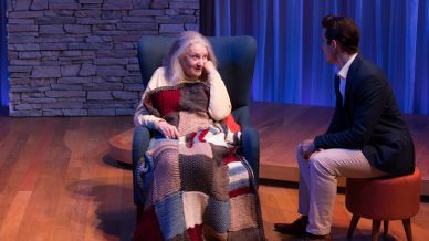 An elderly woman wrapped in a patchwork blanket sits in a blue armchair, looking at a man in a suit who is seated on a small pouffe, with a stone wall and blue-lit curtains in the background.