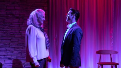 An older woman and a younger man stand facing each other on a stage with pink and purple lighting, next to a wooden chair and curtain backdrop. The woman wears a white blouse and scarf; the man wears a suit jacket.