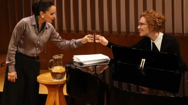 Two women clink glasses and smile at each other; one stands beside a wooden stool with a water jug, while the other sits at a grand piano with sheet music. They are in a warmly lit room with wooden panelling.