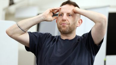A man with short hair and a beard stands indoors, wearing a dark T-shirt and resting both hands on his head, looking slightly upward with a thoughtful or concerned expression.