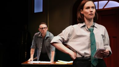 A woman in a striped shirt and green polka-dot tie stands in front of a desk holding a notepad, looking serious. A man stands behind the desk with papers, watching her. The scene appears to be from a theatrical play.